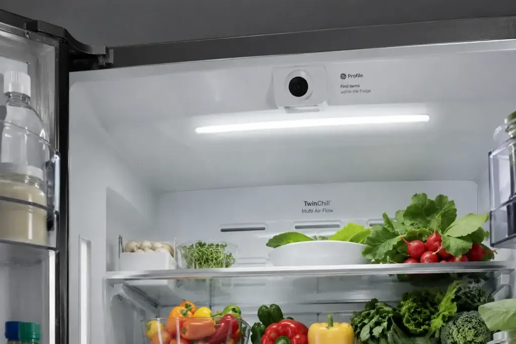 Interior view of a modern GE smart refrigerator with bright LED lighting, an integrated camera module at the top, and fresh vegetables neatly stored on shelves.